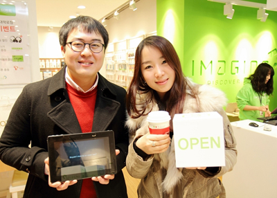 SK플래닛, 카페와 ICT체험 매장을 융합한 ‘이매진(IMAGINE)’ 대학로점 오픈 홍보사진입니다. 왼쪽 모델은 앱 구동화면을 들고 있고 오른쪽 모델은 OPEN이라고 적힌 팻말을 들고 있습니다.