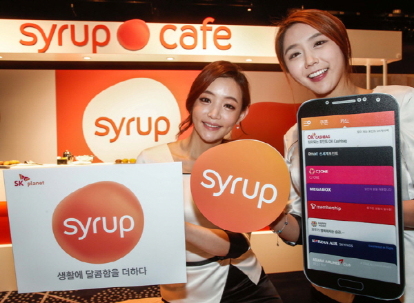 SK플래닛에서 넥스트 커머스를 론칭했다는 홍보사진입니다. 왼쪽 모델은 생활의 달콤함을 더하다 시럽이라고 적힌 배너를 들고 있으며 오른쪽 모델은 앱 아이콘과 핸드폰 앱 구동화면을 보여주고 있습니다.