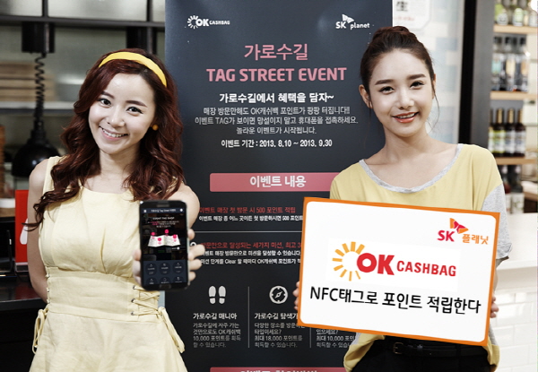OK캐쉬백, NFC태그로 포인트 적립 이벤트 홍보 사진입니다. 왼쪽 모델은 앱 구동화면을 들고 있으며, 오른쪽 모델은 OK캐쉬백 NFC로 포인트 적립한다라고 적힌 배너를 들고 있습니다. 그리고 뒤에 배너에는 가로수길 TAG STREET EVENT라고 적힌 내용과 이벤트 상세 내용이 적혀있습니다.
